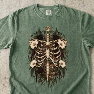 May include: A sage green short-sleeve t-shirt featuring a central graphic print of a human rib cage intertwined with botanical elements including white flowers, ferns, mushrooms, and a moth, topped with a small moon phase illustration.