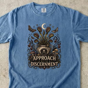 Puede incluir: Una camiseta azul de manga corta con un estampado gráfico de venus atrapamoscas en una maceta, mariposas, setas y una luna creciente. El texto "APPROACH WITH DISCERNMENT" está impreso en una fuente decorativa bajo la ilustración central.