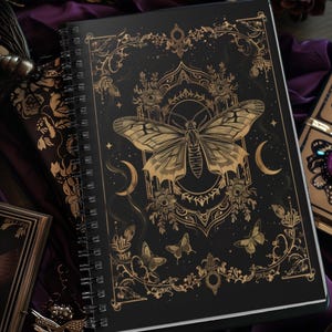 May include: Black spiral-bound notebook with a gold butterfly and floral design. The cover features a large butterfly in the center, surrounded by crescent moons and smaller butterflies. The edges are adorned with gold floral patterns.