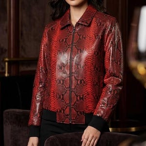 May include: A red snakeskin-effect jacket with a black zip and collar. The jacket has a bomber style with black cuffs and hem. The model is wearing the jacket in a dimly lit room with a dark background.
