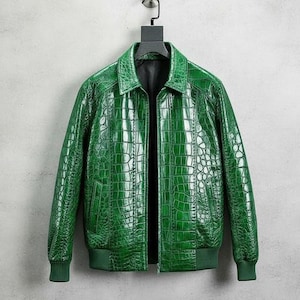 Może przedstawiać: Żakiet ze skóry w żywym zielonym kolorze, o fakturze skóry krokodyla. Kurtka ma kołnierz, zapięcie na zamek błyskawiczny oraz prążkowane mankiety i dół. Wisi na czarnym wieszaku na neutralnym tle.