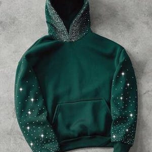 May include: A dark green hoodie with a front pocket and a hood lined with sparkling embellishments. The sleeves are also adorned with scattered, glittering accents. The hoodie is made of a soft material and has a relaxed fit.