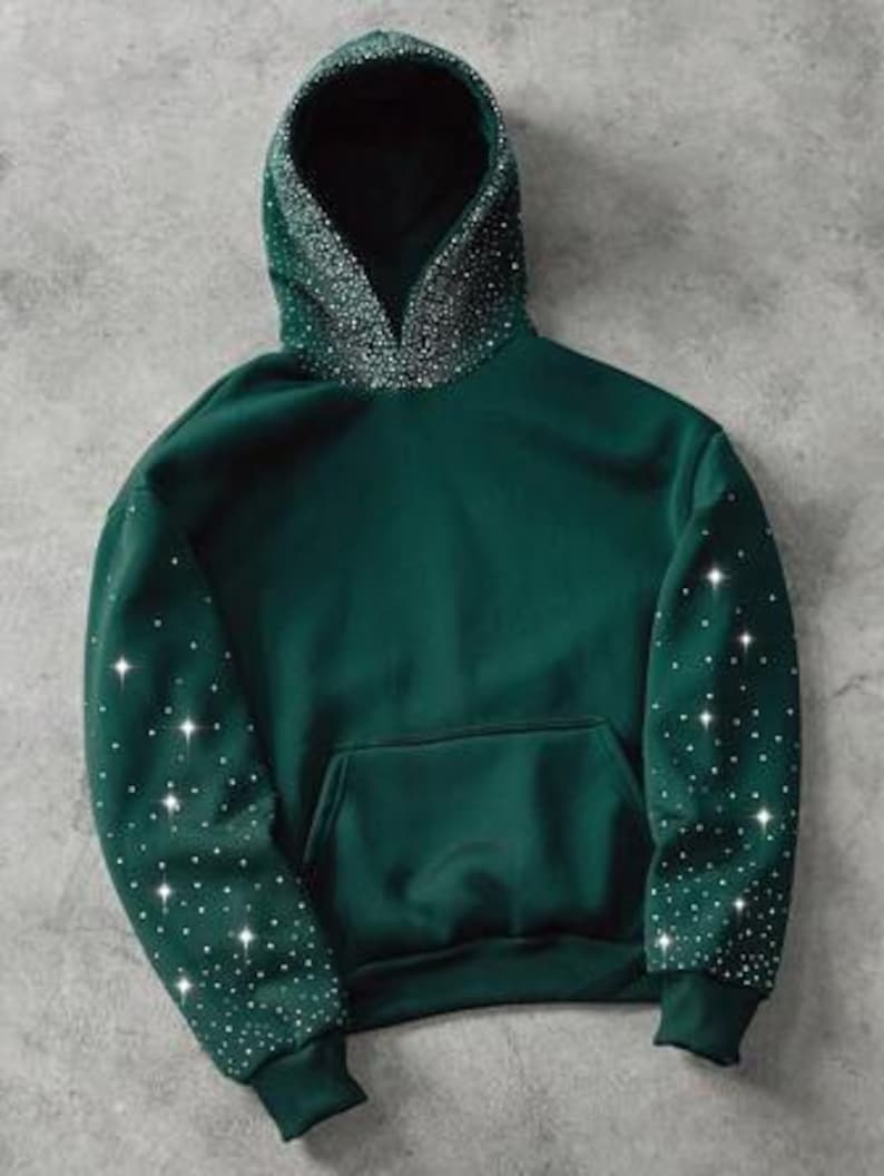 May include: A dark green hoodie with a front pocket and a hood lined with sparkling embellishments. The sleeves are also adorned with scattered, glittering accents. The hoodie is made of a soft material and has a relaxed fit.