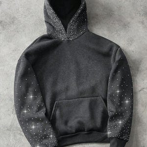 May include: A dark gray hoodie with a front pocket and a drawstring hood. The hood and sleeves are embellished with sparkling white rhinestones. The cuffs and hem are ribbed.