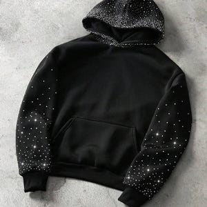 Pode incluir: Moletom preto com bolso frontal e capuz com cordão. O capuz e as mangas são adornados com strass brancos brilhantes. O moletom é feito de um material macio e confortável.