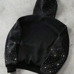 May include: Black hoodie with a hood and long sleeves. The hood and sleeves are embellished with sparkling, scattered rhinestones. The cuffs and hem are solid black.