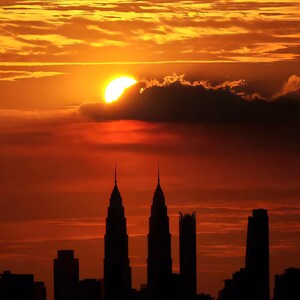 以下が含まれることがあります： 都市のスカイラインに沈む鮮やかな夕日。雲に一部隠れた太陽が、空に温かいオレンジ色の光を投げかけています。シルエットになった建物、特に目立つタワーが、ドラマチックなコントラストを生み出しています。