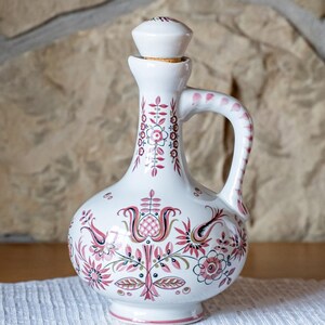 Peut inclure: Une cruche décorative en céramique blanche avec une anse et un bouchon, ornée d'un motif floral rouge. La cruche est posée sur un dessous de plat ovale blanc, devant un mur en pierre.