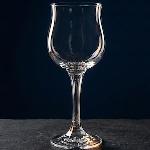 Könnte beinhalten: Ein klares Weinglas mit einer tulpenförmigen Schale und einem langen, schlanken Stiel. Das Glas hat einen runden Fuß und besteht aus transparentem Glas. Der Hintergrund ist dunkelblau.
