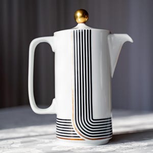 May include: White ceramic coffee press with a gold knob on the lid. The press features black vertical stripes and curved lines with gold accents. The handle is a simple, curved design. A stylish addition to any kitchen.