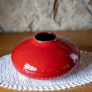 Puede incluir: Un jarrón de cerámica rojo vibrante con una superficie texturizada y un acabado brillante. El jarrón tiene una forma redonda y bulbosa con un interior oscuro y se asienta sobre un tapete de ganchillo blanco. Decoración del hogar.