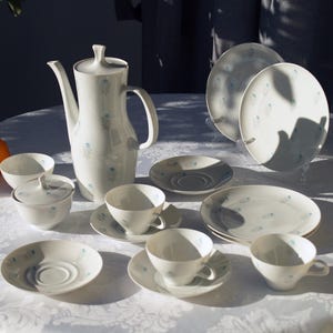 Könnte beinhalten: Ein weißes Porzellan-Teeservice mit floralem Muster, einschließlich einer hohen Teekanne, Tassen, Untertassen, Tellern und einer Zuckerdose. Zwei Orangen liegen auf dem Tisch. Das Set ist auf einer weißen Tischdecke angeordnet.