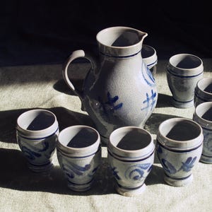 Puede incluir: Una jarra de gres y vasos de chupito a juego. La jarra tiene un asa y un pico, con diseños florales azules. Los vasos de chupito son cilíndricos con detalles azules. El conjunto está dispuesto sobre una superficie clara.