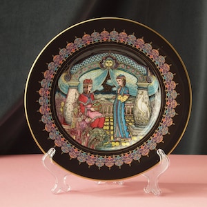 Könnte beinhalten: Dekorative schwarze Platte mit goldenem Rand und farbenfrohem Rand. Die Platte zeigt eine detaillierte Illustration von zwei Figuren in einem nahöstlichen Setting. Die Platte wird auf einem transparenten Kunststoffständer präsentiert.