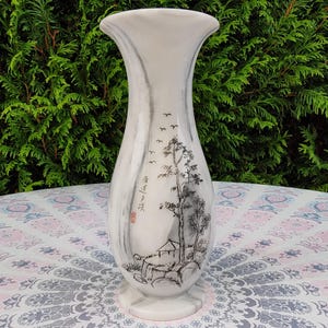 Puede incluir: Jarrón de cerámica blanca con la parte superior acampanada y base ancha. El jarrón presenta un diseño de paisaje en blanco y negro con árboles, una pequeña casa y pájaros volando. El jarrón tiene una textura similar al mármol y es adecuado para la decoración del hogar.