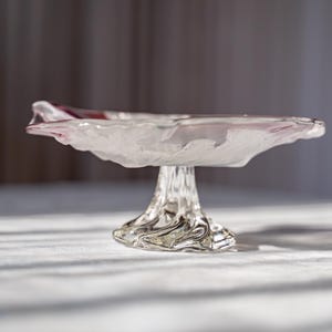 Könnte beinhalten: Eine transparente Glasschale mit Fuß und strukturiertem, organischem Design. Die Schale hat einen ausgestellten, blütenblattartigen Rand mit einem Hauch von Rosa. Der Sockel ist ein detailliertes, geformtes Design. Ein dekoratives Stück zum Servieren oder Ausstellen.
