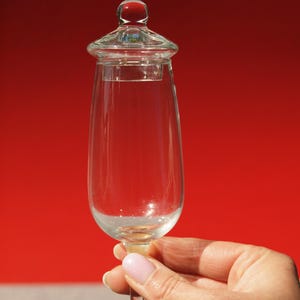 Könnte beinhalten: Ein klares Glasbecher mit Deckel, gefüllt mit einer klaren Flüssigkeit. Der Becher wird von einer Hand vor rotem Hintergrund gehalten. Der Deckel hat einen kleinen roten Knopf.