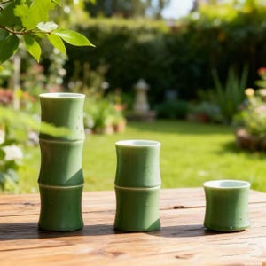 Puede incluir: Tres tazas de cerámica verde, diseñadas para parecerse a tallos de bambú, se colocan sobre una superficie de madera. Las tazas tienen un acabado brillante y un interior verde claro. El fondo presenta un jardín verde borroso.