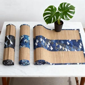 Peut inclure: Chemin de table en bambou avec des accents de tissu à motifs bleus et blancs. Le chemin de table est enroulé en trois sections, dont une entièrement déployée. Une plante en pot avec de grandes feuilles vertes est posée sur la table.