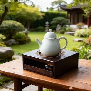 Op de afbeelding: Een witte keramische theepot met deksel staat op een donkere houten theeverwarmer. De verwarmer heeft een lade en een klein metalen kopje. De opstelling staat op een houten tafel, met een tuin op de achtergrond.