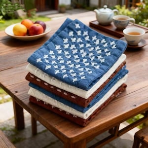 May include: A stack of square cloth napkins in blue, white, and brown. The top napkin is blue with white embroidered patterns. The napkins are stacked on a wooden table, with a bowl of fruit and a tea set in the background.
