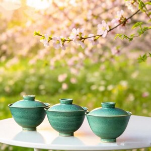 May include: Three teal-colored ceramic bowls with lids sit on a white table. The bowls have a rounded shape and a glossy finish. The background features a blurred view of a flowering tree with pink blossoms, suggesting a spring theme.