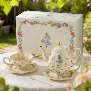 Könnte beinhalten: Cremefarbenes Teeservice zum Thema Alice im Wunderland. Das Set enthält eine Teekanne, eine Teetasse, eine Untertasse und einen kleinen Löffel. Die Teekanne ist auf einer Teetasse und Untertasse gestapelt. Die Box zeigt Illustrationen von Alice und Blumendesigns.