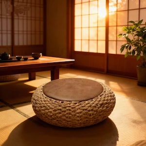 Puede incluir: Un otomán redondo tejido con un cojín marrón. El otomán está colocado sobre una estera de tatami en una habitación con una mesa de madera y puertas correderas japonesas tradicionales. La habitación está iluminada por la luz del sol.