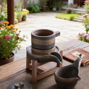 May include: A ceramic tea set with a cup, a stand with a spout, a small pitcher, and a lid. The set is in shades of brown and gray, with a rustic, handcrafted appearance. The set is placed on a wooden table outdoors.