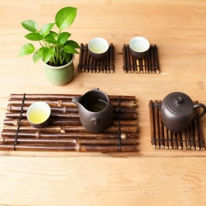 Peut inclure: Une composition de service à thé avec une petite théière marron, une verseuse et deux tasses, le tout reposant sur des nattes en bambou. Une petite plante en pot avec des feuilles vertes ajoute une touche naturelle.