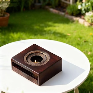 May include: A dark brown wooden candle holder with a decorative brass ring around the candle. The candle holder is square with a small drawer at the bottom. The candle flame is lit, casting a warm glow. The candle holder is on a white table.