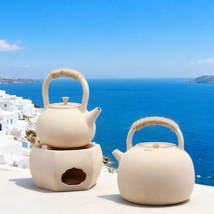 Puede incluir: Dos teteras de cerámica color crema con asas envueltas en cuerda. Una tetera se encuentra sobre un calentador a juego con una abertura decorativa. Las teteras están sobre un fondo de mar y cielo azules, con edificios blancos a lo lejos.