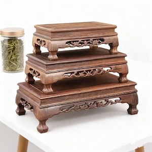 Może przedstawiać: Trzy ułożone w stos, ciemnobrązowe drewniane stojaki ekspozycyjne z ozdobnymi rzeźbionymi detalami. Każdy stojak ma prostokątny blat i dekoracyjne nogi. Po lewej stronie znajduje się szklany słoik ze złotą pokrywką i zieloną zawartością.