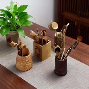 May include: A collection of wooden tea ceremony tools, including brushes, scoops, and tongs, displayed in bamboo and woven containers. A potted green plant sits in the background. The tools are arranged on a gray table runner.