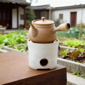 Puede incluir: Una tetera de cerámica marrón con tapa, colocada sobre un calentador de cerámica blanco. El calentador tiene una abertura circular con un borde dorado. El asa de la tetera está envuelta con hilo. El conjunto está sobre una superficie de madera, con un jardín al fondo.