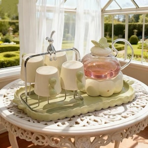 Peut inclure: Service à thé vert pastel comprenant une théière en verre et des tasses assorties. La théière a un couvercle en forme de papillon et les tasses ont des poignées vertes arrondies. Un lapin argenté est assis sur le porte-tasses. Le thé est rose.