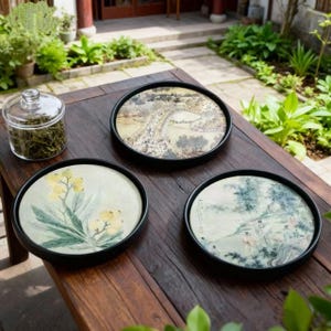 Op de afbeelding: Drie ronde decoratieve trays met zwarte randen, elk met een ander aquarelontwerp. Eén tray toont een landschap, een andere toont gele bloemen en de derde toont een serene scène. Een glazen pot met theebladeren staat in de buurt.