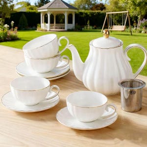 Puede incluir: Un juego de té de porcelana blanca con ribete dorado, que incluye una tetera, tazas, platillos y un infusor de té. La tetera tiene un adorno dorado. El juego está dispuesto sobre una superficie de madera, con un jardín y un cenador al fondo.