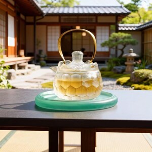May include: A clear glass teapot with a gold handle, filled with tea, sits on a green glass saucer. The teapot has a unique honeycomb design. The background features a traditional Japanese garden and architecture.