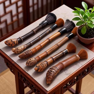 Puede incluir: Seis pinceles de caligrafía con mangos de bambú y extremos tallados están dispuestos sobre una mesa. Los cabezales de los pinceles son negros y marrones. Los mangos varían en color de marrón claro a oscuro. Una pequeña planta en maceta está en el fondo.