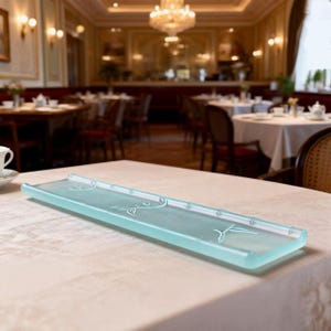 Puede incluir: Una bandeja rectangular de vidrio azul claro con un diseño de bambú. La bandeja es larga y estrecha, con bordes elevados y detalles de tallos y hojas de bambú. La bandeja está sobre un mantel blanco.