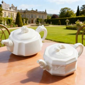 May include: Two white porcelain teapots with a floral design on a wooden table. The teapots have a unique shape and are set against a backdrop of a large house and a green lawn.