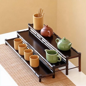 Puede incluir: Una exhibición de juego de té de bambú en niveles. Los estantes de color marrón oscuro sostienen tazas de bambú, un soporte para utensilios de bambú y teteras y una crema de cerámica en tonos marrones y verdes.