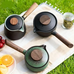 May include: Three ceramic teapots with wooden handles and lids. One is black, one is green, and one is black with a light green interior. The teapots are on a white cloth, with a jar of tea leaves and fruit nearby.