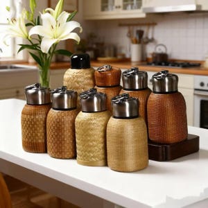 May include: A collection of woven containers with stainless steel lids and a black lid. The containers are in various shades of brown and beige, with a textured, basket-like appearance. They are arranged on a white surface, with a kitchen in the background.