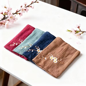 Peut inclure: Quatre serviettes en lin pliées, rouges, turquoise, bleu marine et marron. Chaque serviette est ornée d'une broderie florale blanche. Elles sont disposées sur une surface blanche, avec des fleurs roses en arrière-plan.