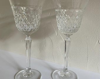 May include: Two clear crystal wine glasses with diamond-cut patterns on the bowls. The glasses have long, faceted stems and round bases. The background is a plain white surface.