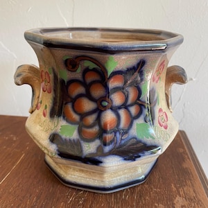 May include: A decorative ceramic planter with a hexagonal shape and two handles. The planter features a large floral design in shades of orange, blue, and green, with smaller pink flowers. The background is a beige color with a crackle finish.