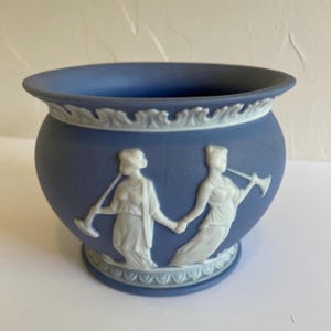 Könnte beinhalten: Eine blaue Keramikschale mit weißen Relief-Figuren von Frauen, die Instrumente spielen. Die Schale hat eine runde Form mit einer weiten Öffnung und einem dekorativen Rand. Der Boden der Schale ist ebenfalls mit einem weißen Reliefmuster verziert.