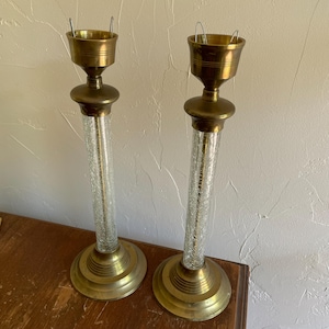 May include: Two vintage brass candle holders with clear crackle glass columns. The candle holders have a round base and a cup-shaped top to hold the candles. The brass has a warm, aged patina.